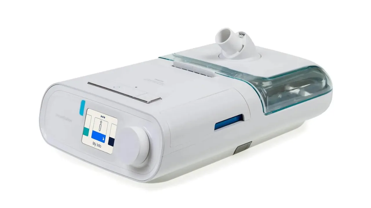 CPAP Philips DreamStation branco visto de três quartos, com a tela na frente, o botão giratório ao lado e a câmara de água transparente do umidificador acoplada à direita